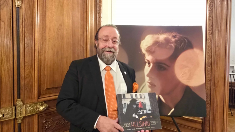 Jussi Brofeldt mit der Publikation AHO &amp; SOLDAN – Helsinki 1950-luvun varjossa, vor einer Fotografie von Claire Aho während des Empfangs in Wien. Foto: CulturaLatina.
